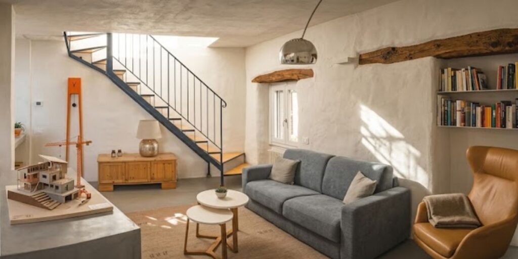 Modern living room with a grey sofa, tan armchair, floating stairs, bookshelves, and rustic wooden beams.