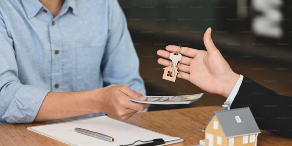 Two people exchange a hundred-dollar bill for a house key over a wooden table with a small model house nearby.